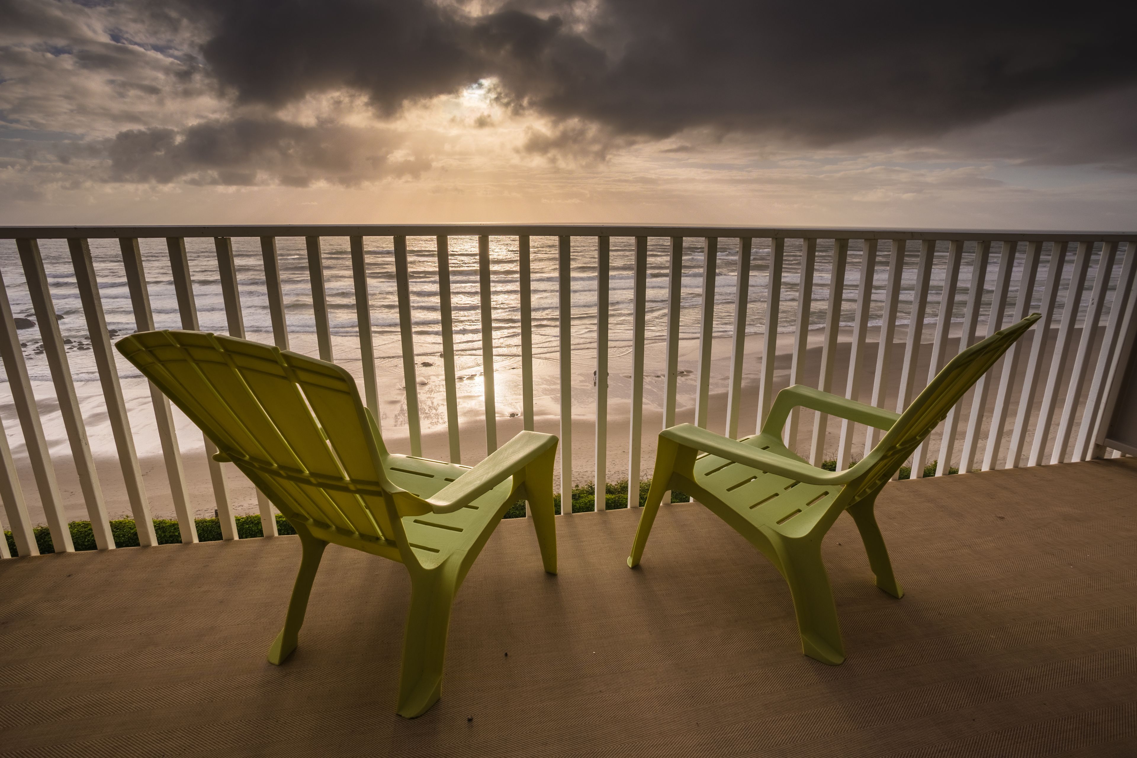 Two green chairs on a wooden deck overlooking the ocean with a white railing.
