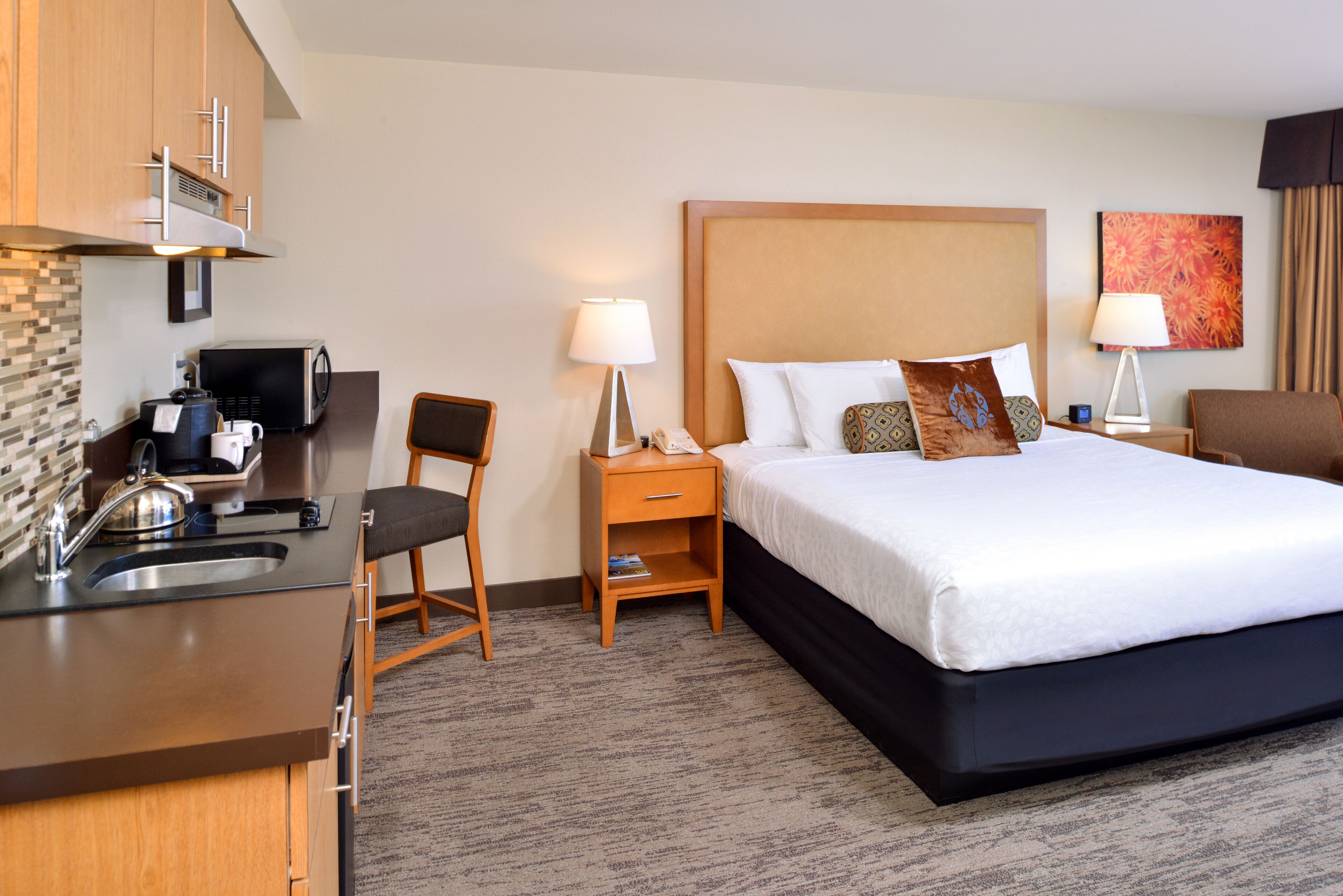 A hotel room with a bed, bedside table, lamp, chair, and kitchenette.