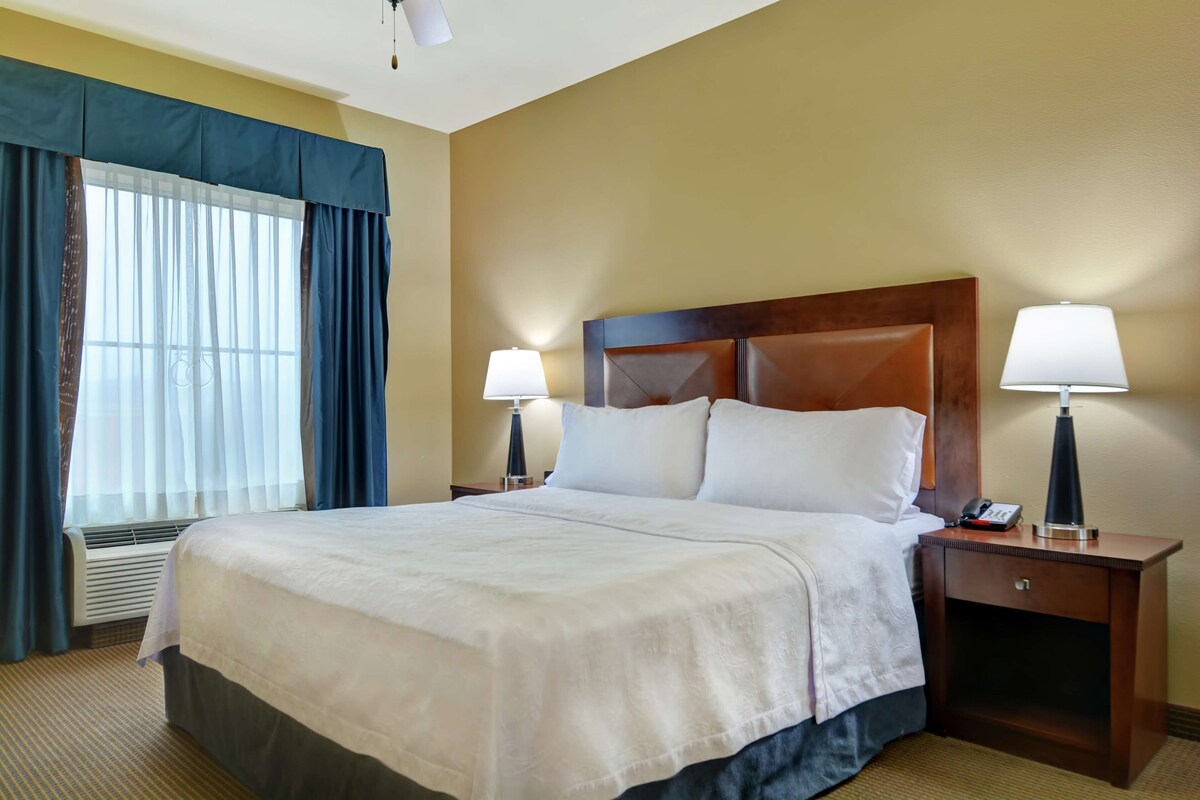 A hotel room with a large bed, two bedside tables with lamps, a nightstand, and a window with curtains.