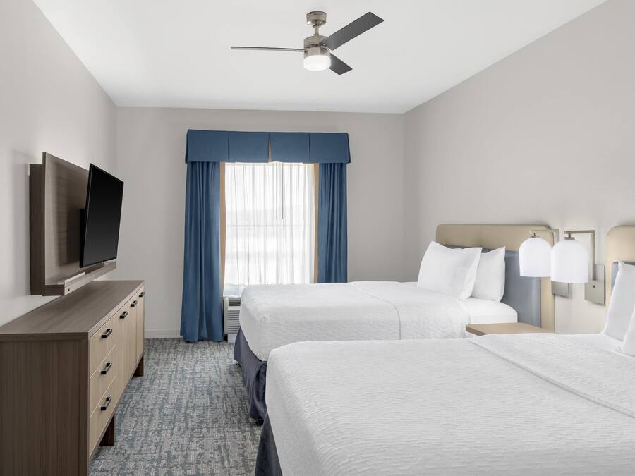 A hotel room with two beds, a TV on a wooden dresser, a ceiling fan, and a window with blue curtains.