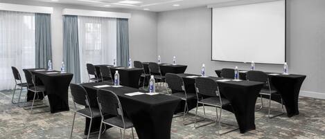 Un salón de conferencias con mesas, sillas, una pantalla de proyector y botellas de agua.