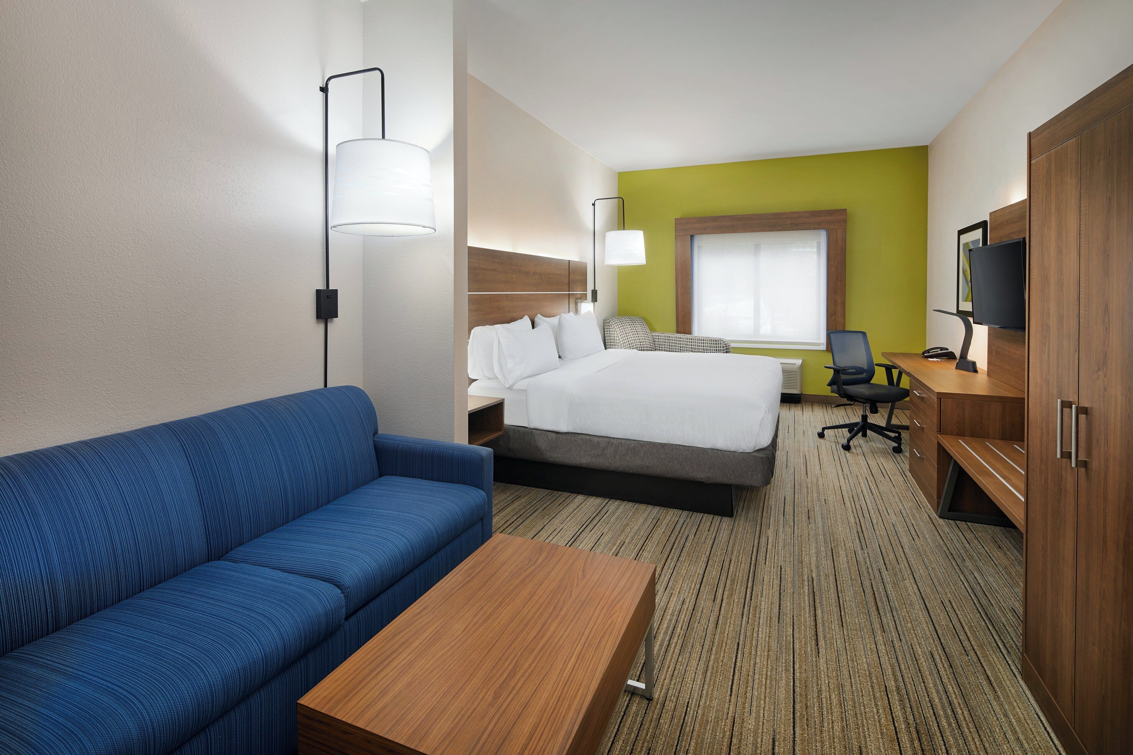 A hotel room with a bed, a blue sofa, a wooden desk, a television, and a window.