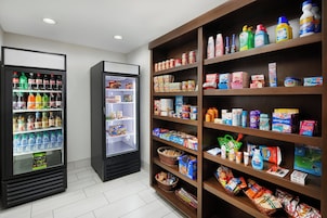 A self-service beverage area with multiple refrigerators stocked with drinks and a shelf with various food and household items.