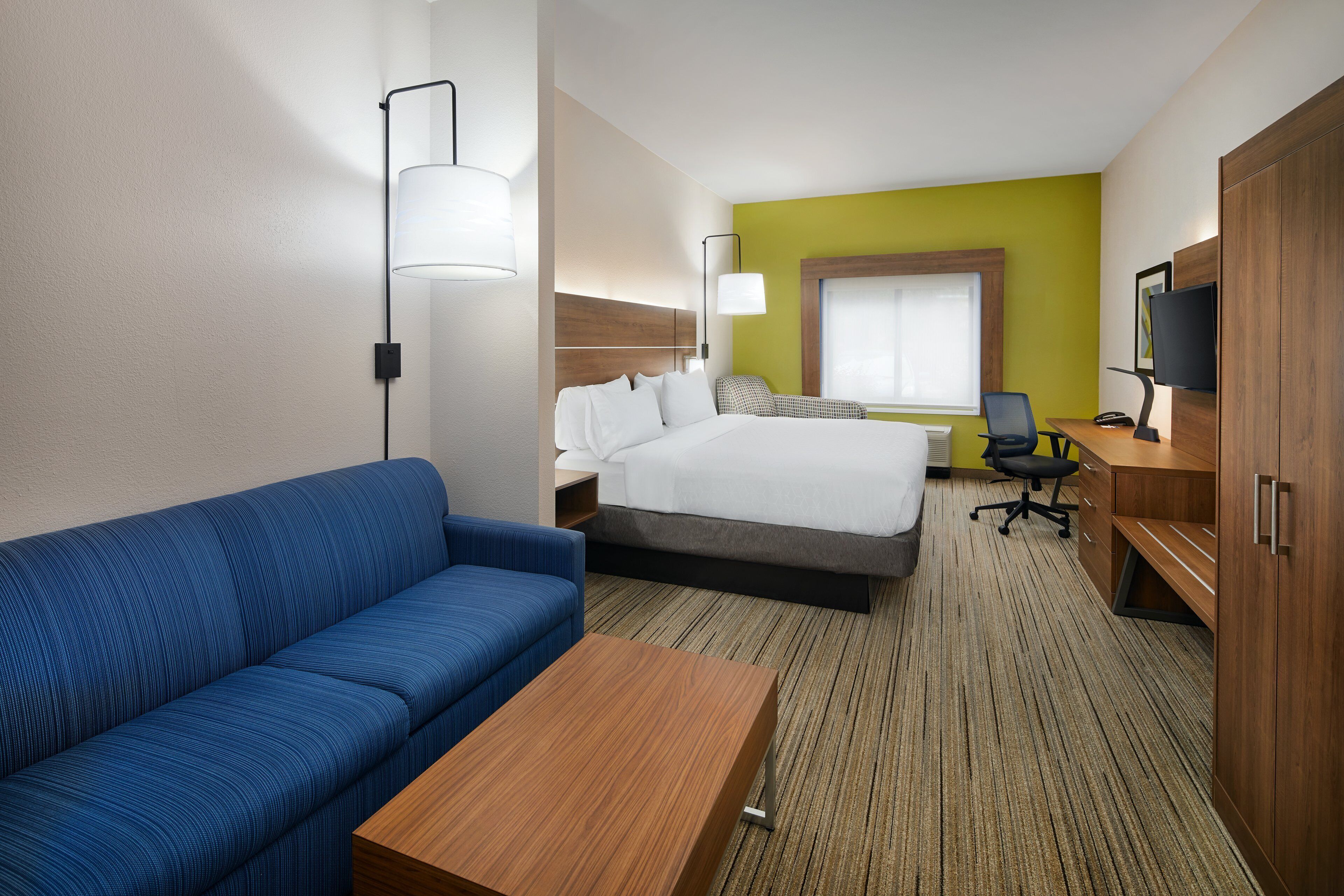 A hotel room with a bed, a blue sofa, a wooden desk, a TV, and a window with blinds.