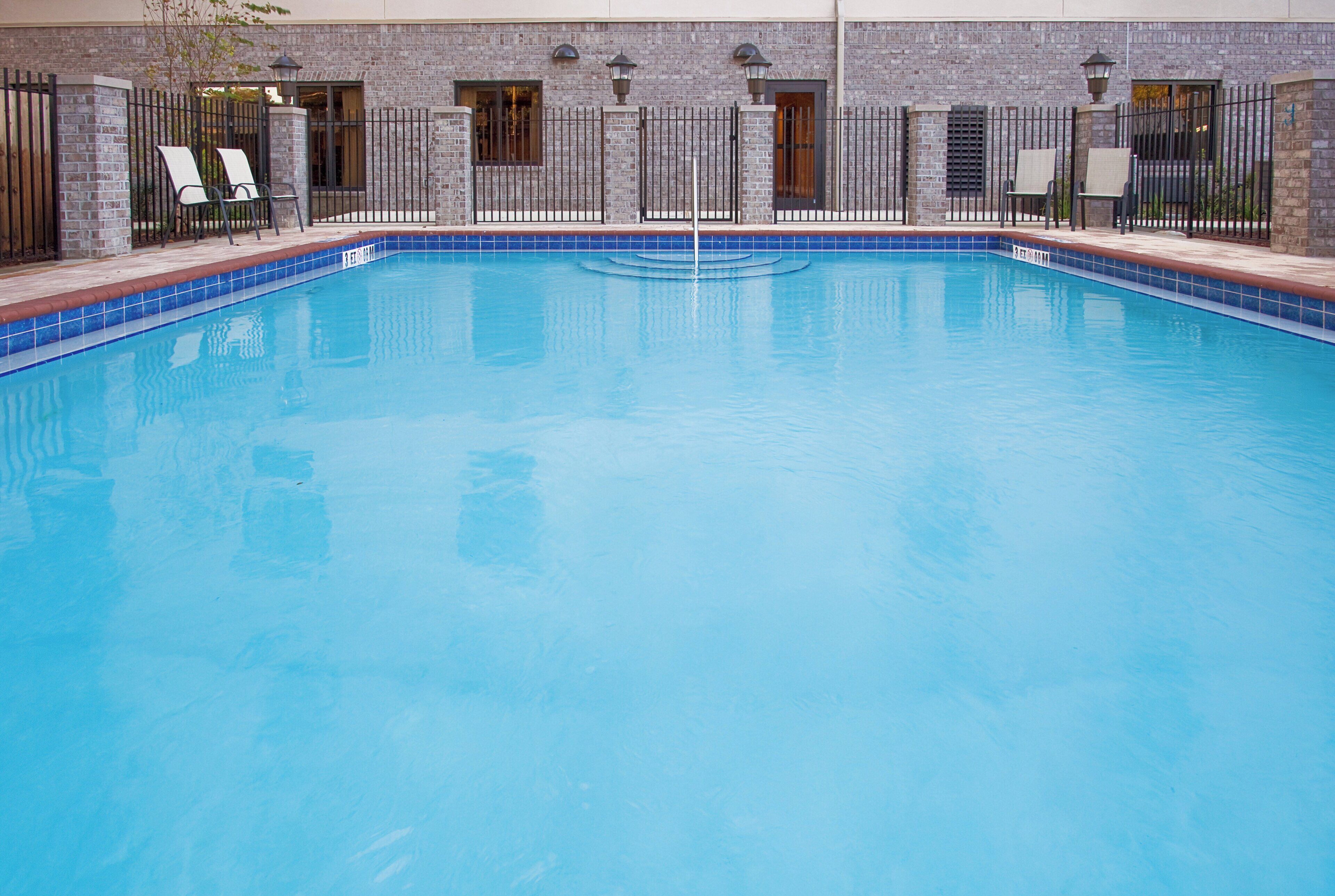 A rectangular indoor swimming pool with a tiled edge, surrounded by a brick wall with windows and a metal fence.