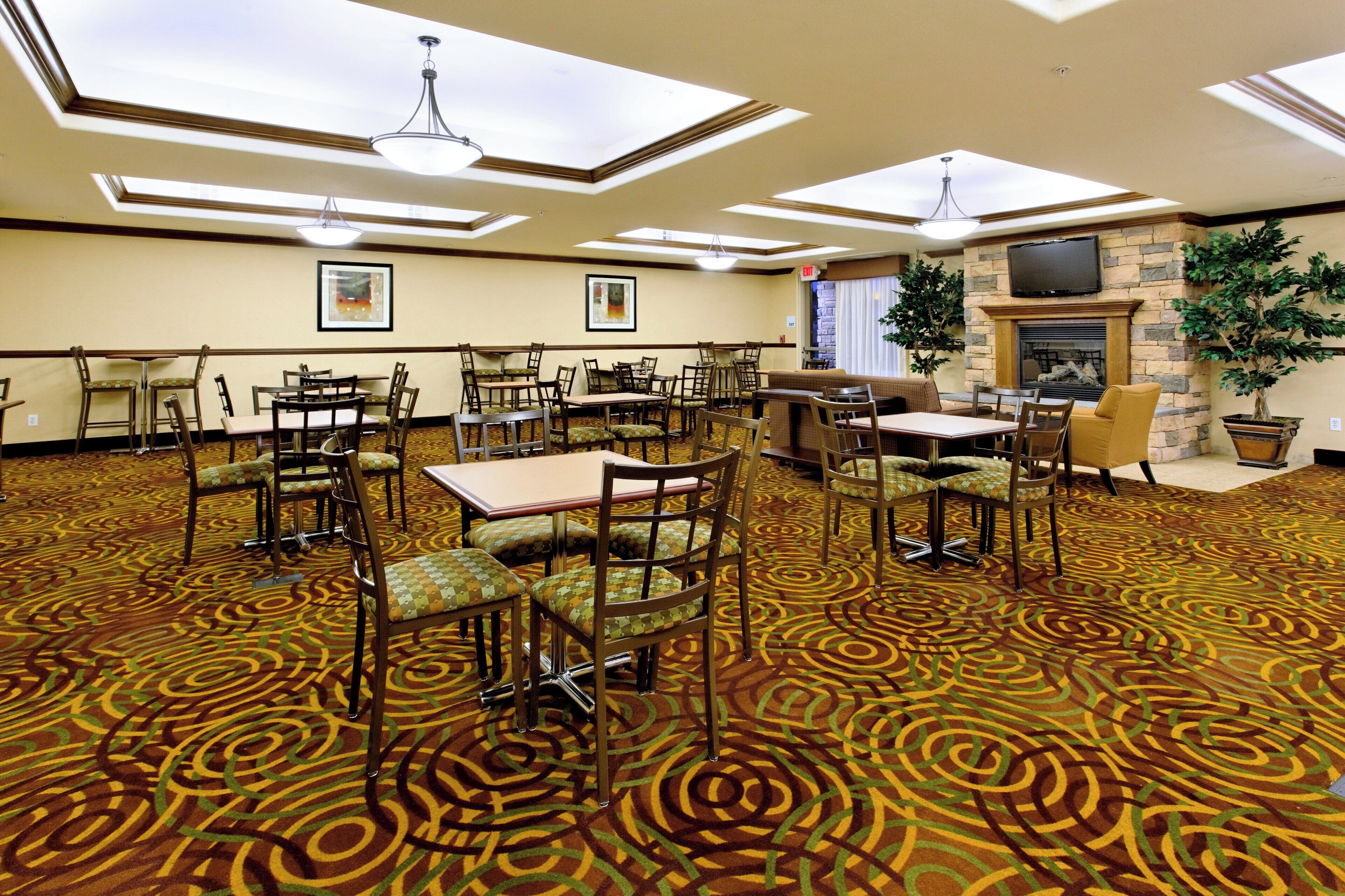 A hotel dining area with multiple tables and chairs, a fireplace, and a large plant.
