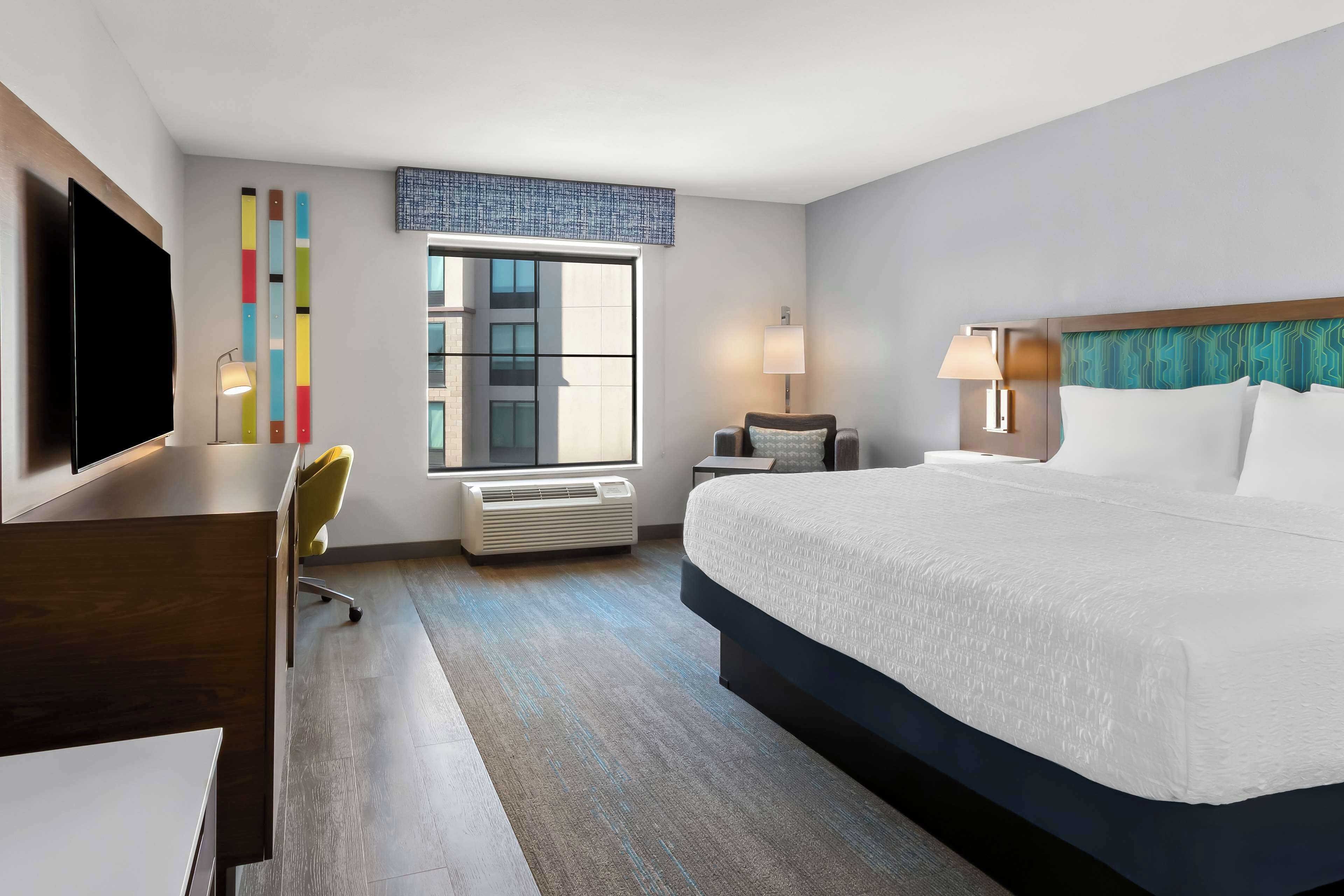 Une chambre d’hôtel avec un grand lit, un bureau, une chaise, une télévision, une fenêtre et un mur orné de rayures verticales colorées.