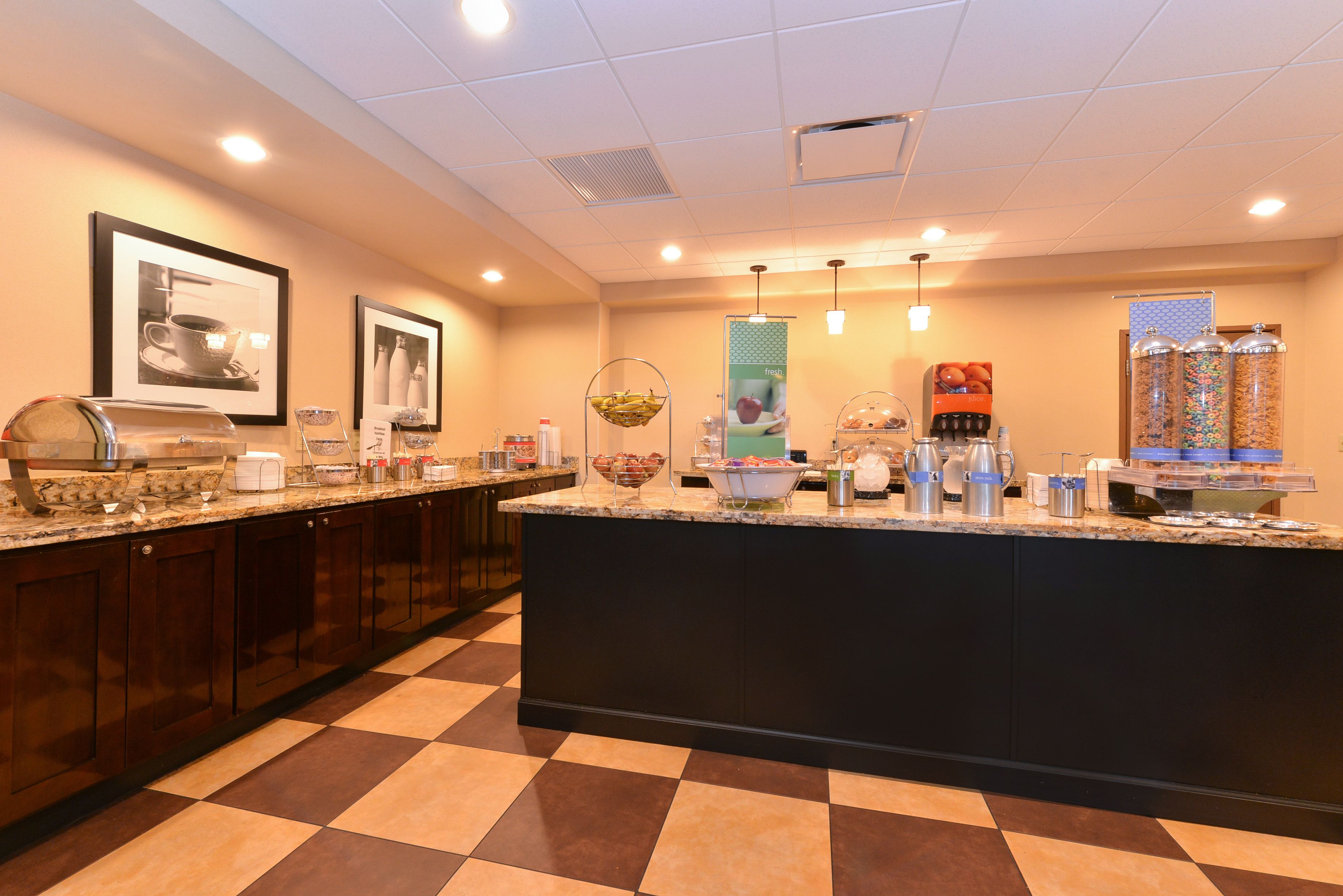 A hotel breakfast area with a checkered floor, a central island with food and drink dispensers, framed pictures on the wall, and a large mirror.