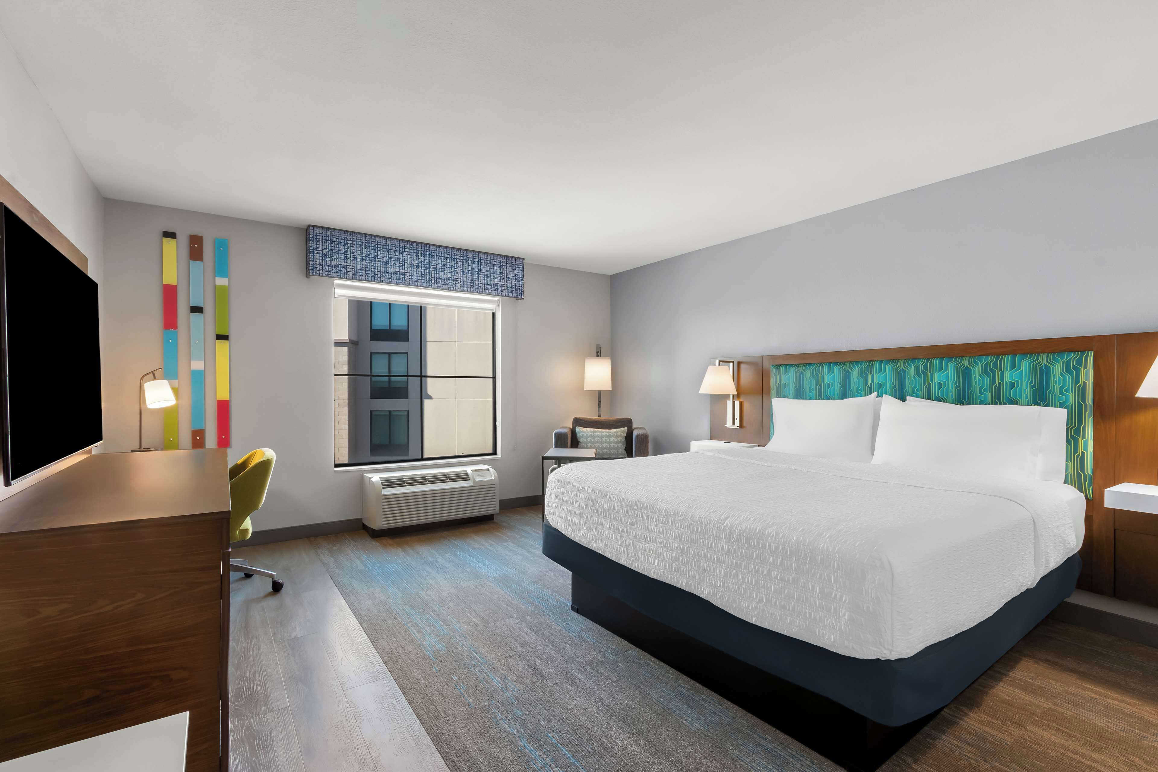 A modern hotel room with a large bed, a desk, a chair, a television, and a window with blinds.