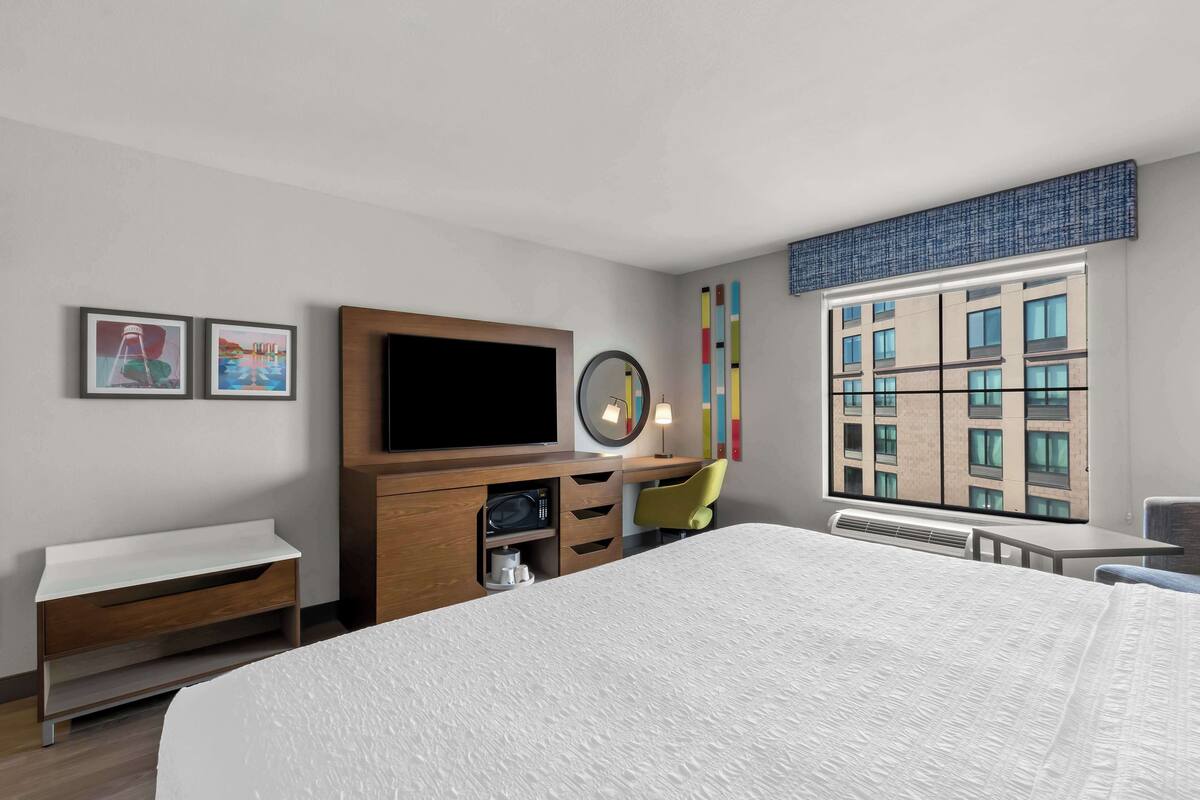 Une chambre d’hôtel avec un grand lit, une télévision, un bureau, une chaise et une fenêtre donnant sur des immeubles.