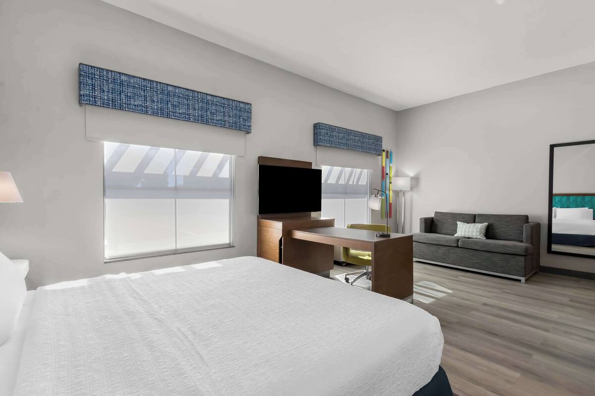 Une chambre d’hôtel moderne dotée d’un grand lit, d’un bureau, d’une télévision, d’un canapé et de grandes fenêtres.