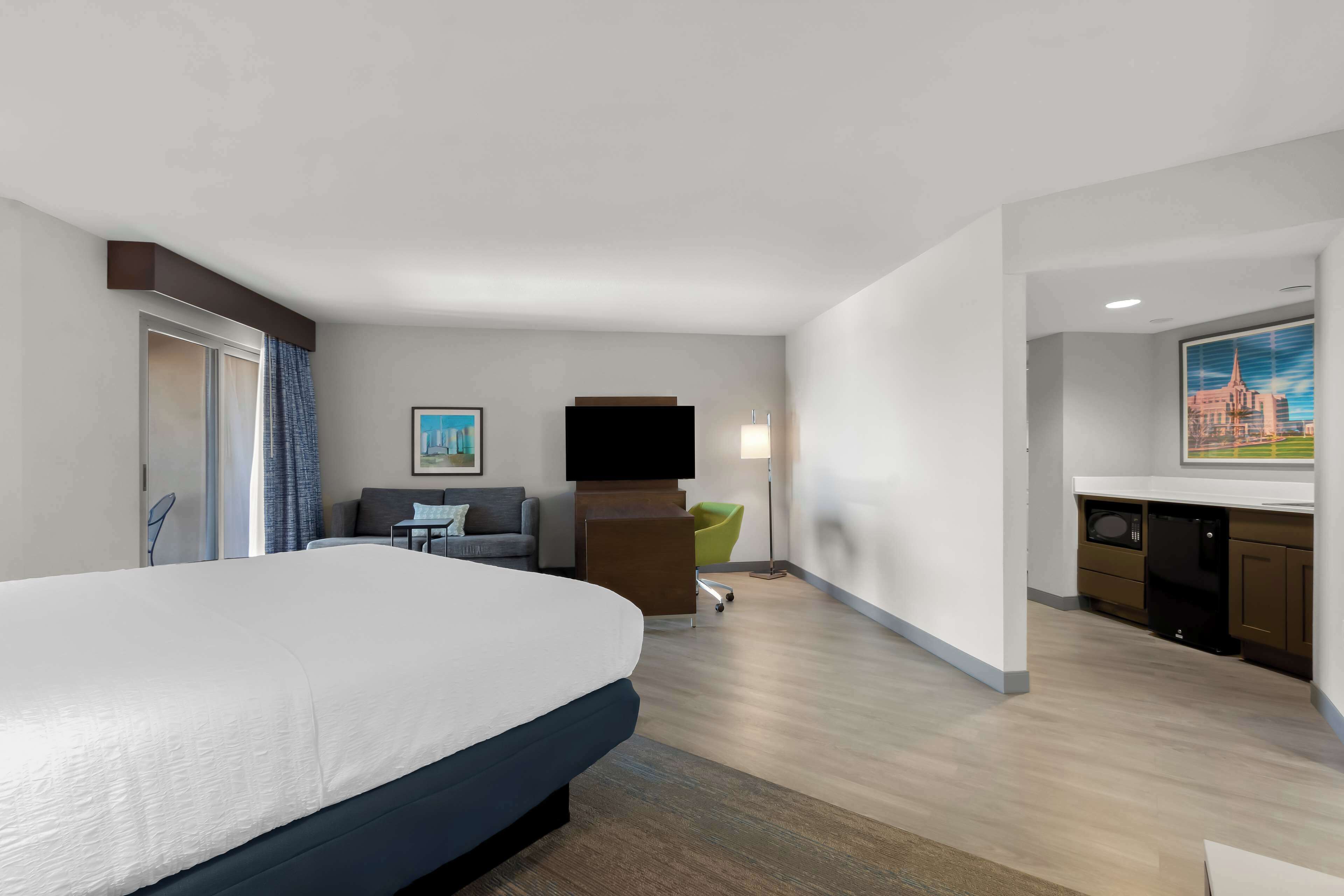 A hotel room with a large bed, a desk, a chair, a flat-screen TV, and a kitchenette.