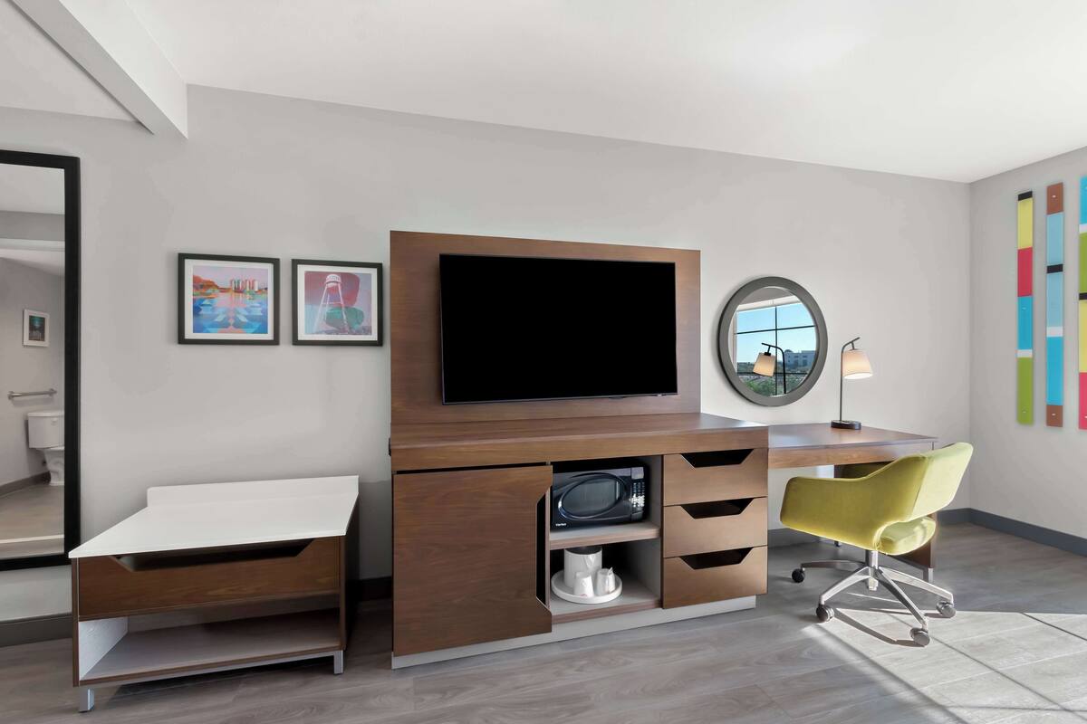 Une chambre moderne avec un bureau en bois, une télévision, une chaise jaune et des œuvres d’art accrochées aux murs.