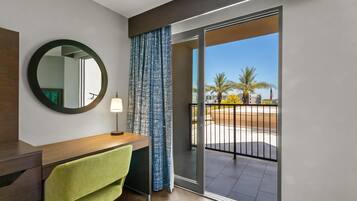 A modern hotel room with a wooden desk, a green towel, a round mirror, and a sliding glass door leading to a balcony with a view of palm trees and buildings.