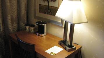 A hotel room with a wooden desk, a lamp, a framed picture of a tree, and a trash bin.
