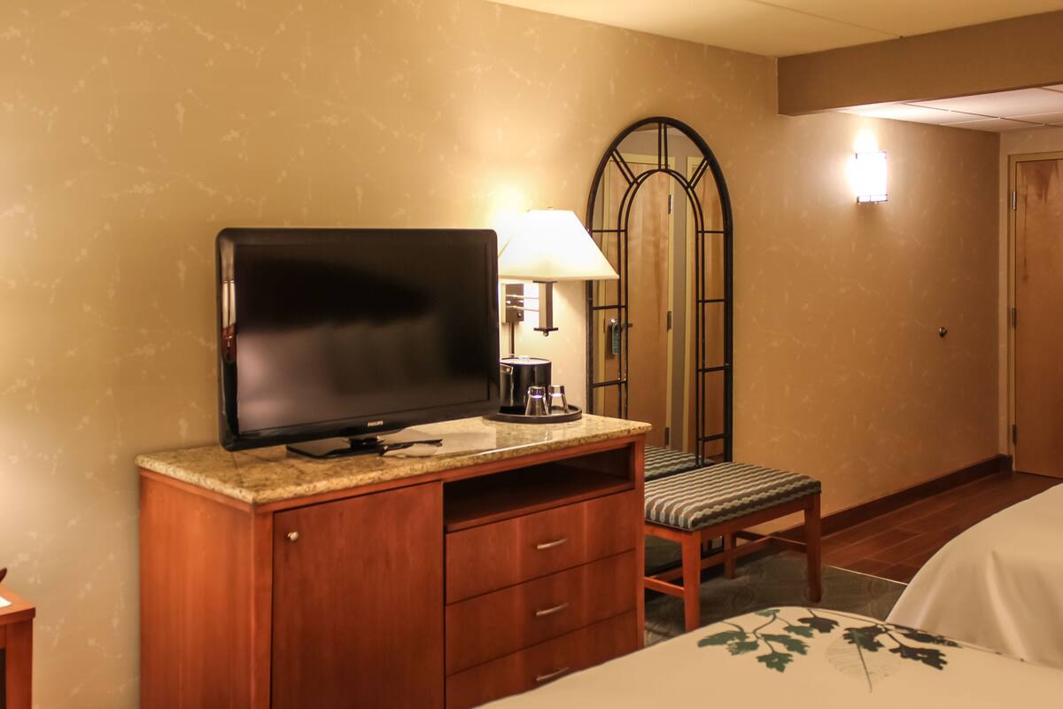 A hotel room with a television on a wooden cabinet, a chair, a desk with a lamp, and a bed with a floral bedspread.