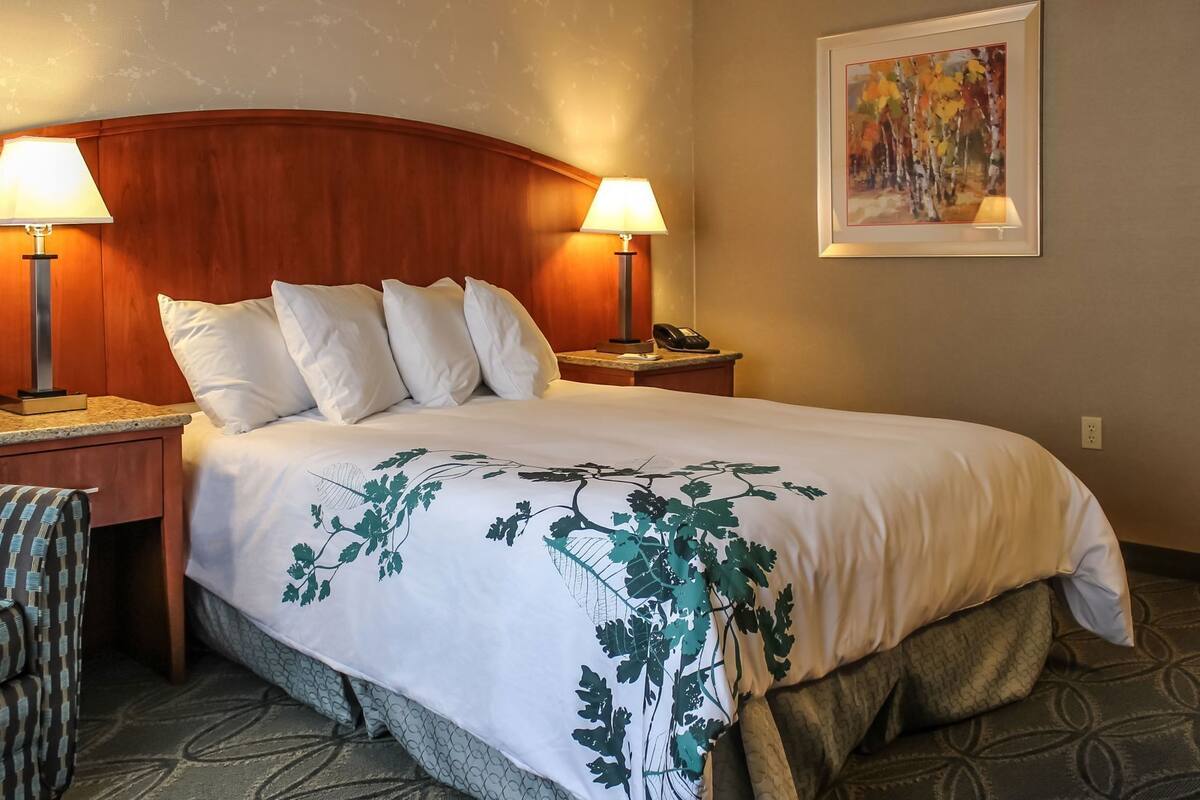 A hotel room with a large bed, two bedside lamps, a telephone, a chair, and a painting on the wall.
