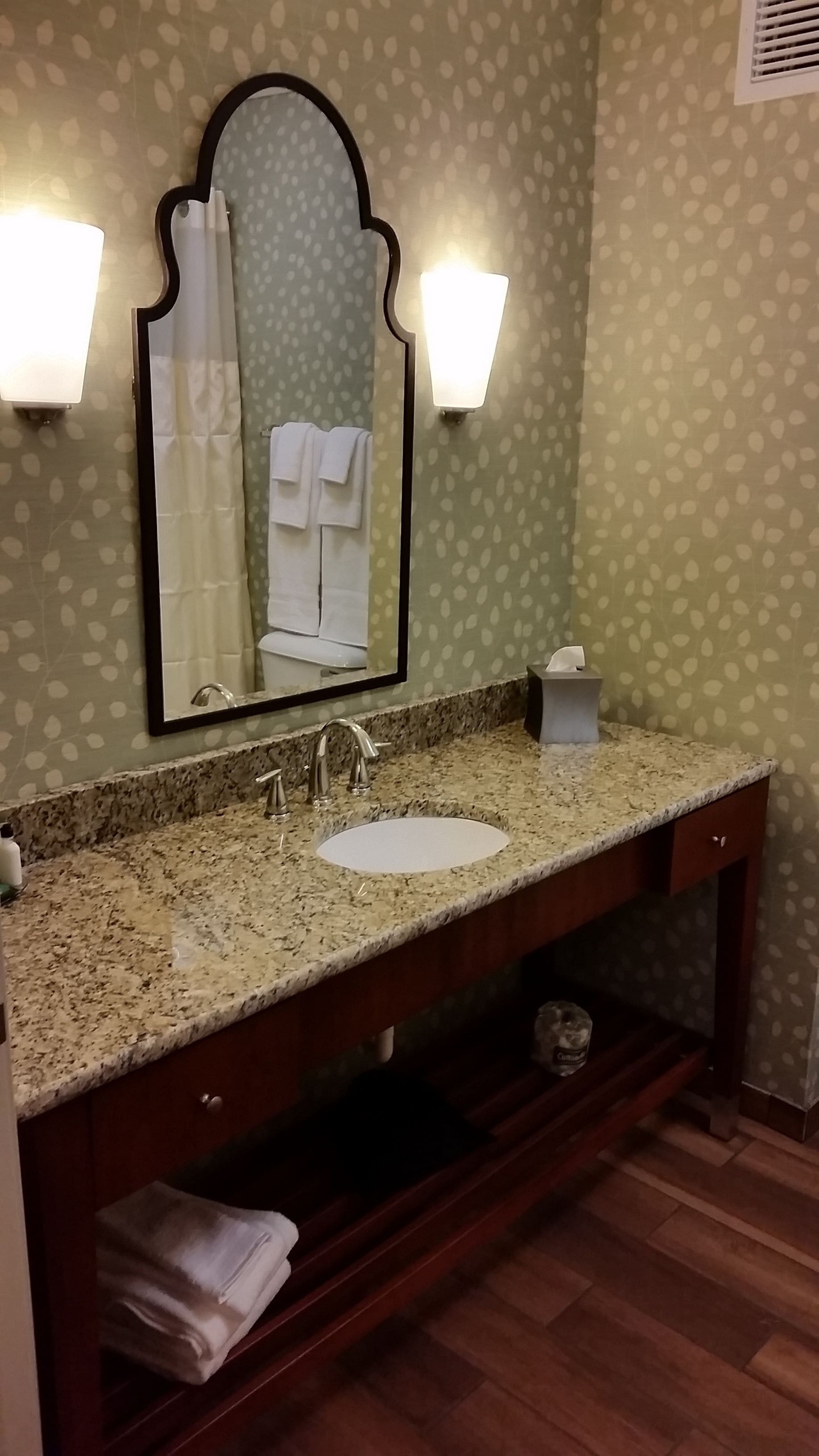 A bathroom with a granite countertop sink, a mirror with a decorative frame, and two wall-mounted lights.
