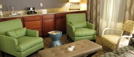 Een hotelkamer met een kitchenette, twee groene fauteuils, een beige poef en een gestreept sofa.