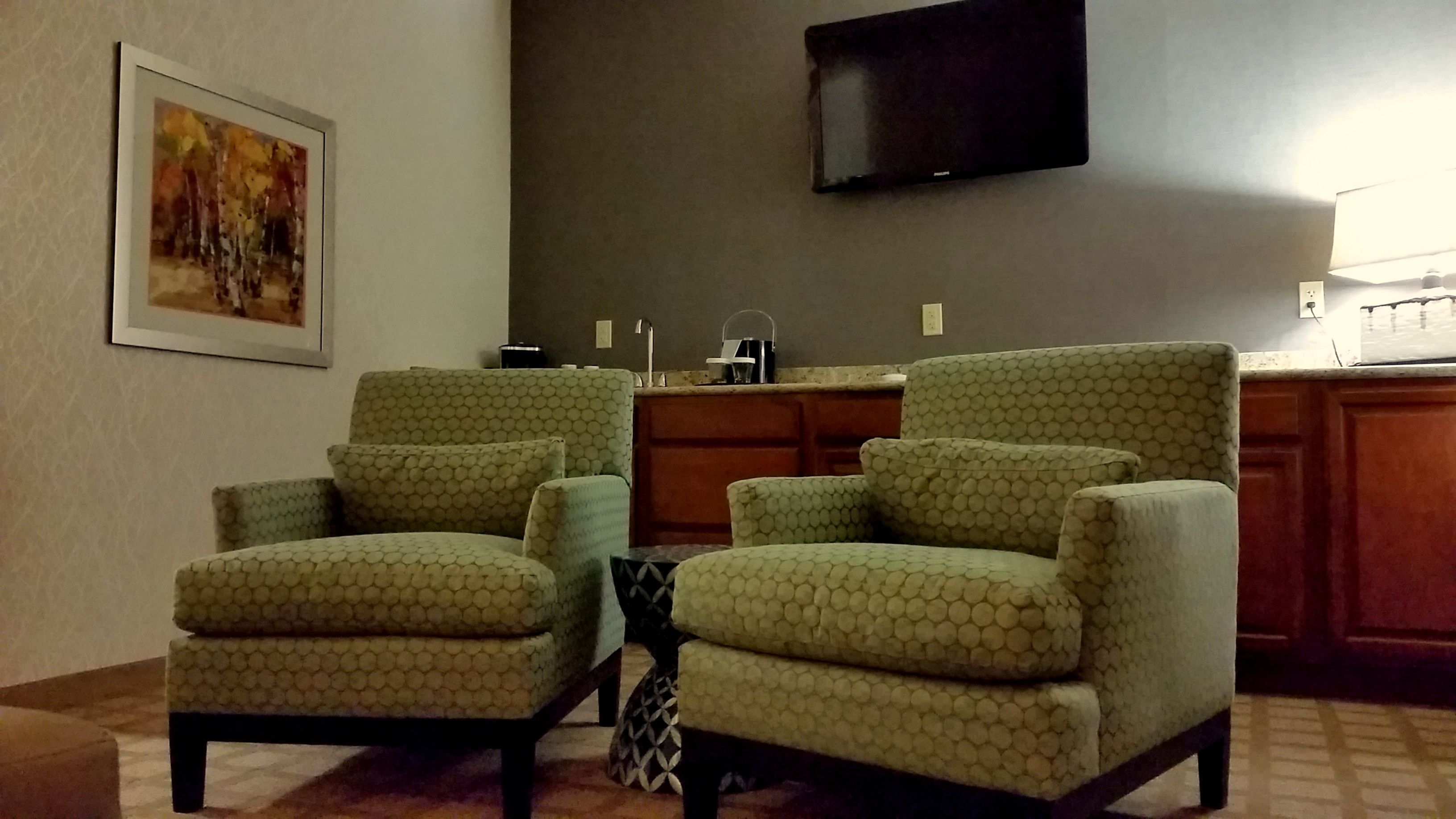 A living room with a sofa set, a painting on the wall, and a flat-screen TV mounted above a kitchen area.