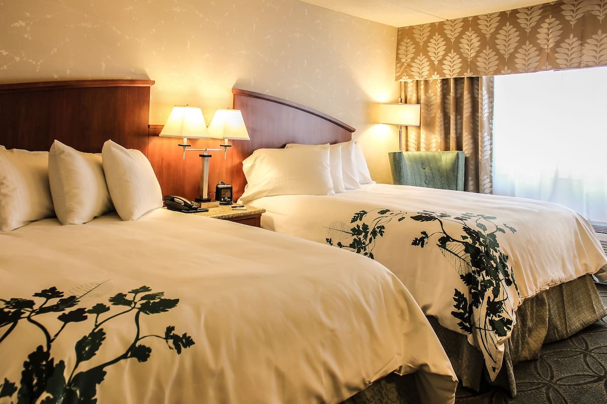 A hotel room with two beds, a nightstand, lamps, and a view of the city.