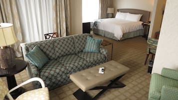 Quarto de hotel com sofá, mesa pequena com luminária, cama e uma cadeira.