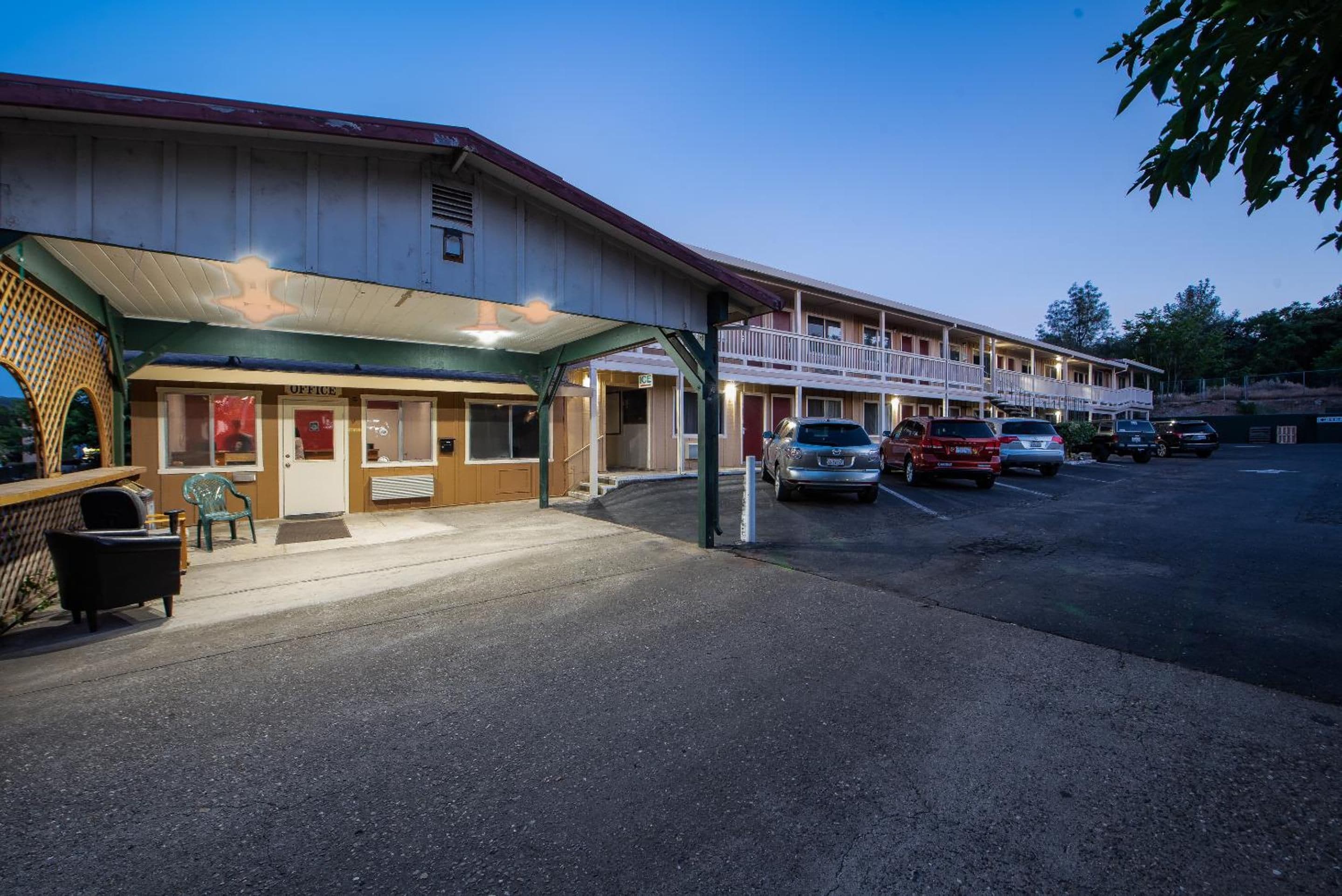 Um motel com área frontal coberta, várias vagas de estacionamento e uma fileira de quartos com varandas.