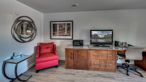 A compact office space with a red chair, a wooden desk with a computer and a monitor, a framed picture on the wall, and a round mirror. - Angels Inn (Angels Camp)