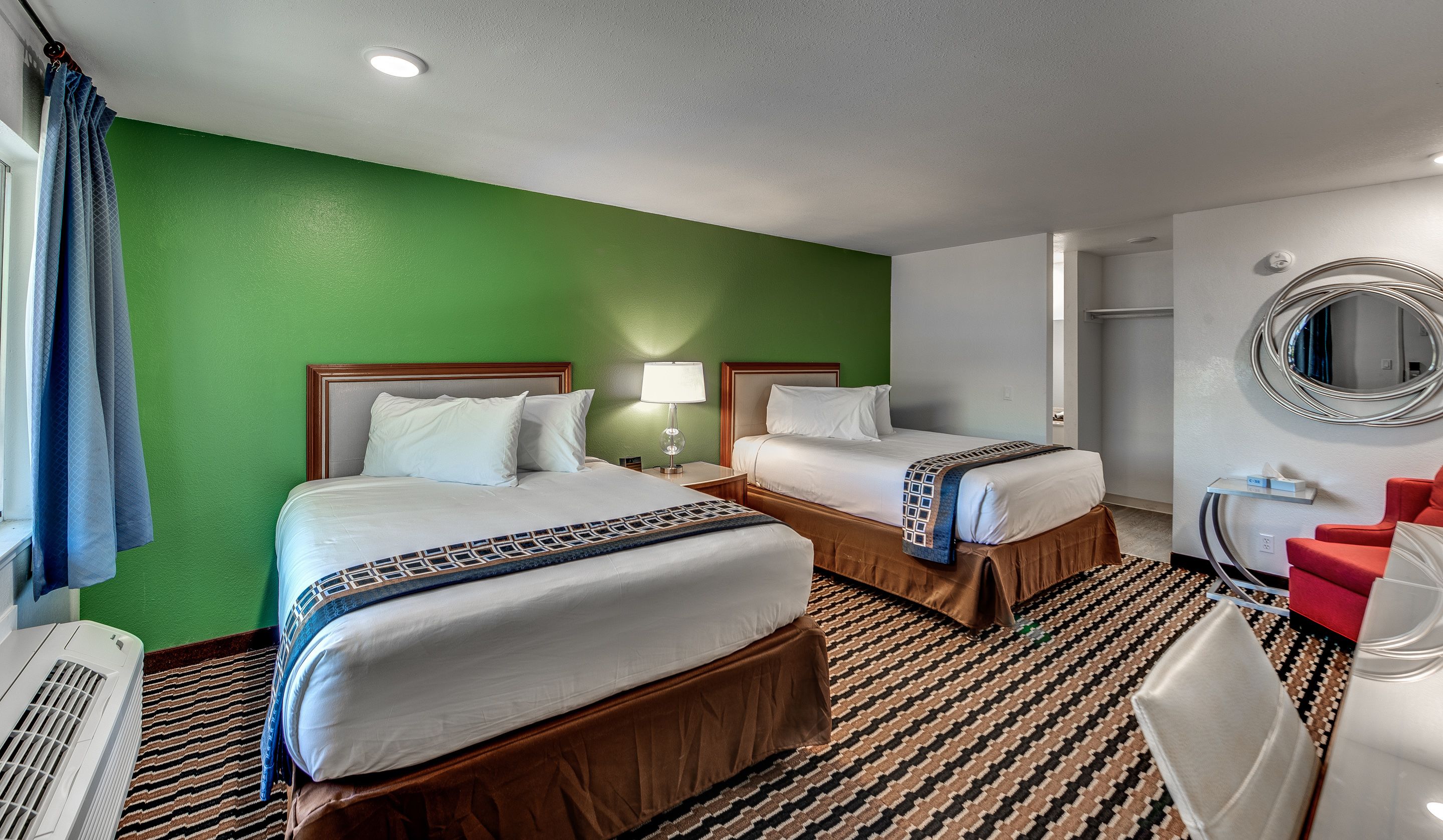A hotel room with two beds, a green wall, a patterned carpet, a red chair, and a round mirror on the wall.