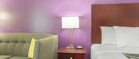 Een hotelkamer met een groene bank, een klein houten nachtkastje met een lamp en een wit bed.