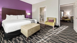 Habitación de hotel con cama, sillón, reposapiés, alfombra estampada y un acceso a un baño.