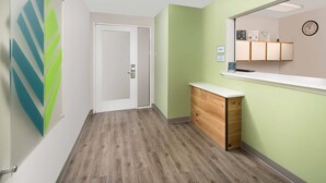 A hallway with a wooden floor, a white door, and a reception desk with a mirror and clock. - WoodSpring Suites Spartanburg - Duncan (Duncan)