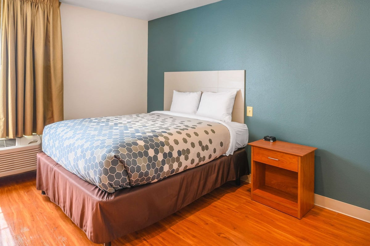 A neatly made bed with a patterned bedspread and pillows, a wooden nightstand with a drawer, and a window with curtains.