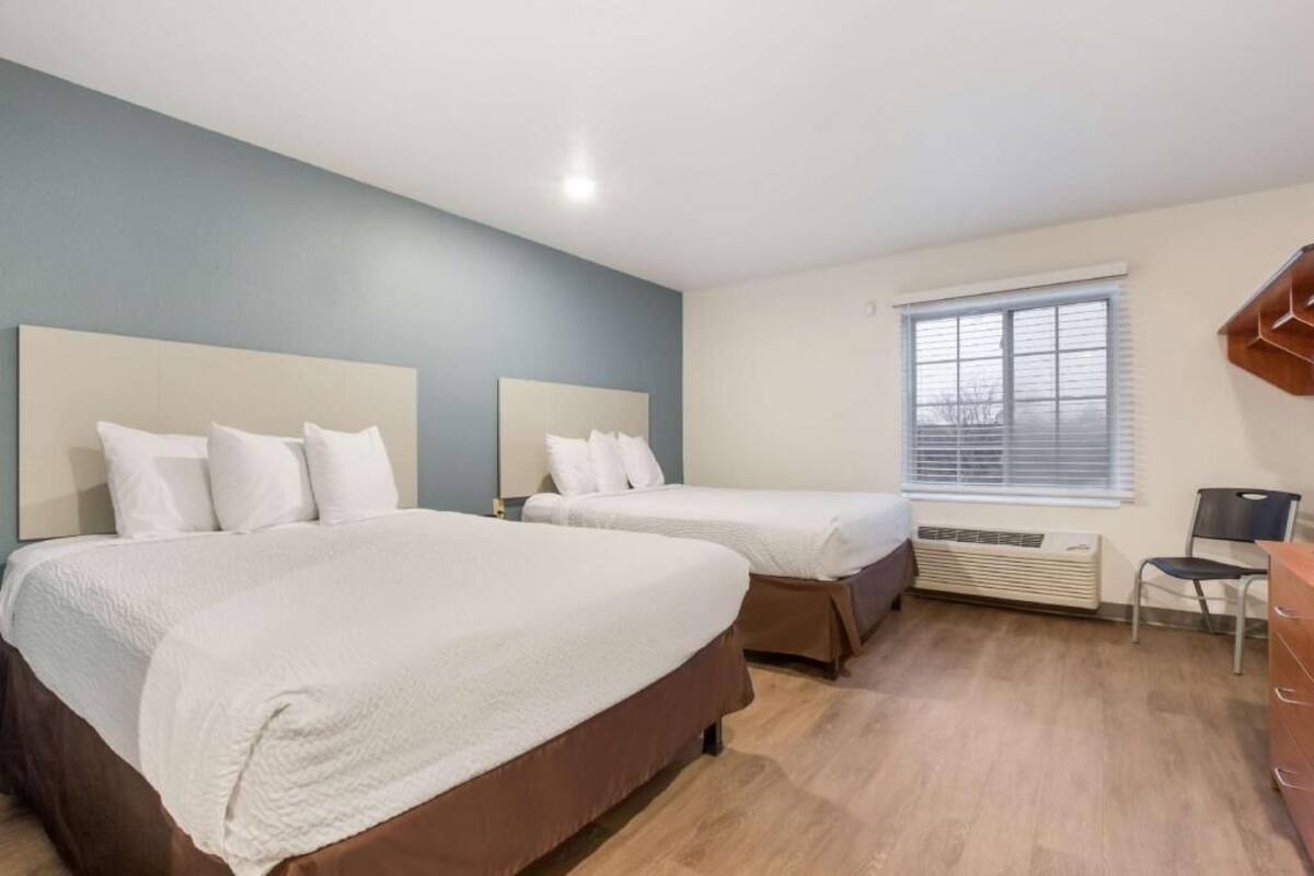 Quarto de hotel com duas camas, uma janela, ar condicionado e uma escrivaninha com cadeira.