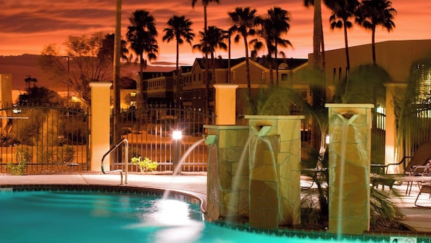 Une piscine avec un ciel au coucher du soleil, des palmiers et un bâtiment en arrière-plan.
