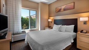A hotel room with a large bed, a flat-screen TV, a heater, a bedside table with a lamp, and a window with curtains.