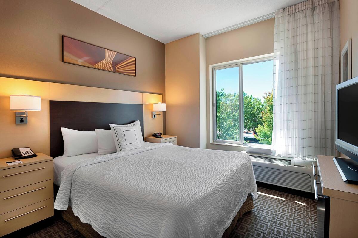 A hotel room with a large bed, two bedside tables with lamps, a flat-screen TV, and a window with curtains.