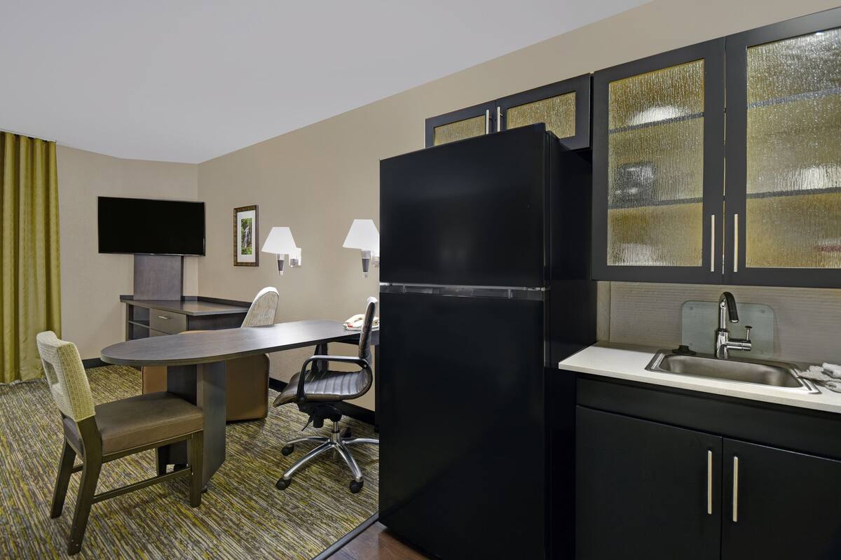 Habitación de hotel compacta con kitchenette, un televisor de pantalla plana montado en la pared, una mesa de comedor con dos sillas y una pequeña zona de trabajo.