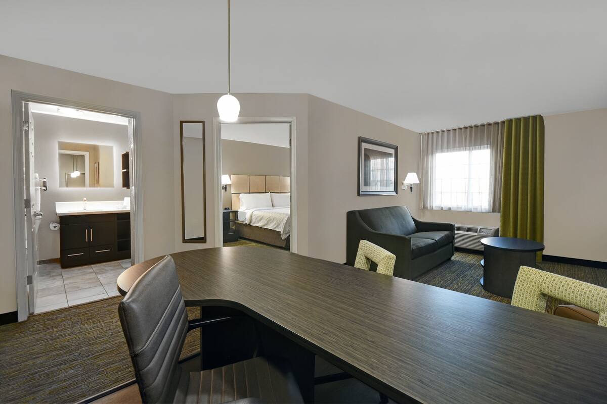 Habitación de hotel con comedor, cama, baño y un amplio ventanal con cortinas.