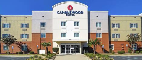 A multi-story hotel with a prominent 'CANDLEWOOD SUITES' sign, a parking lot, and landscaped areas with small trees and shrubs.