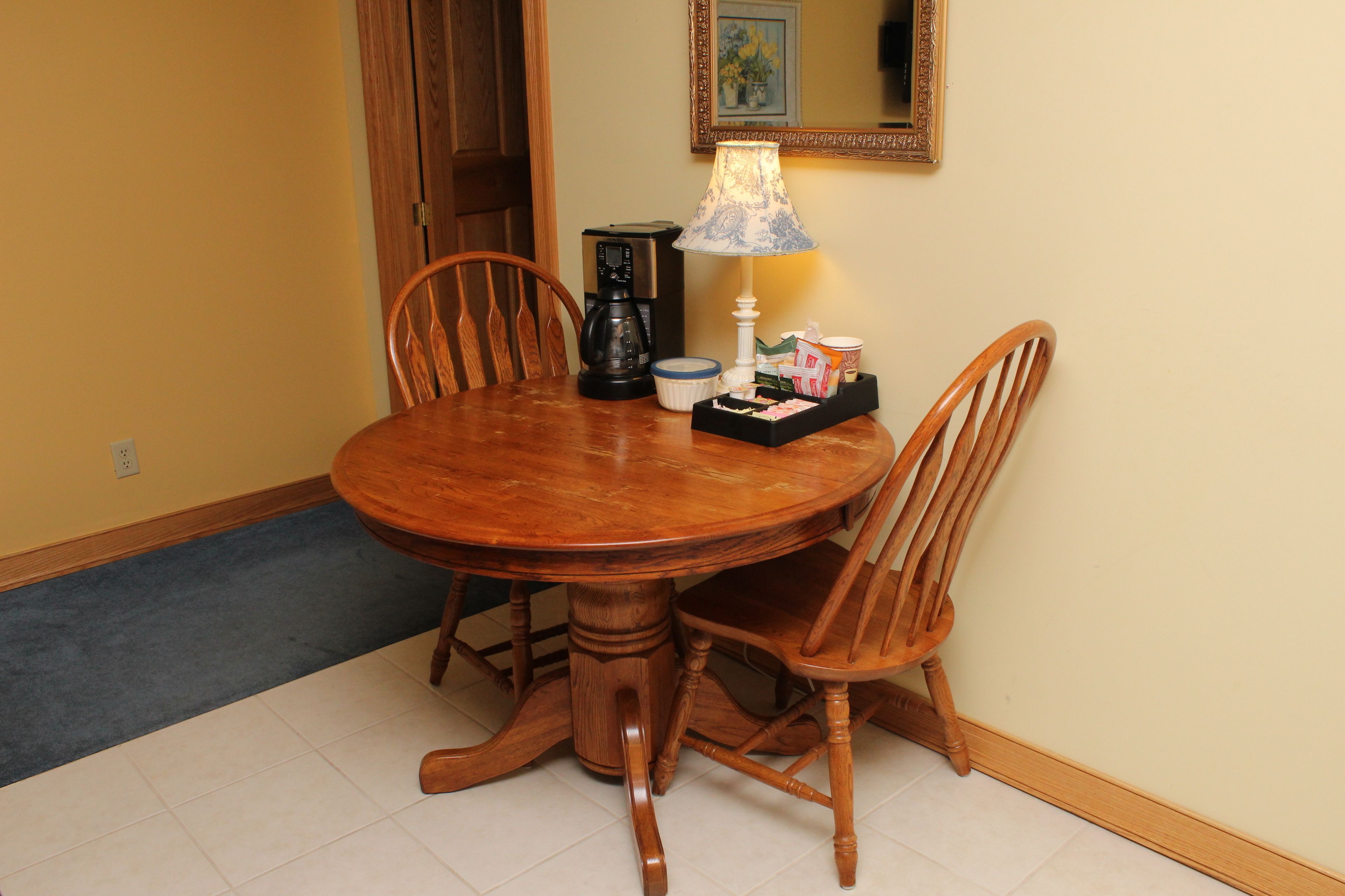 Una mesa de madera para comer con cuatro sillas, una cafetera, una lámpara y una bandeja con salsas y aderezos.