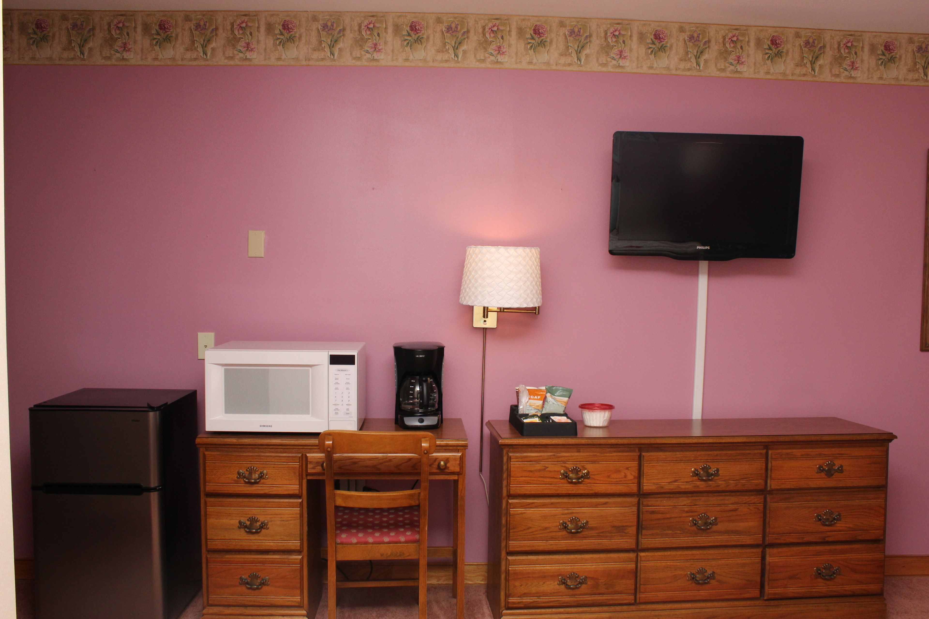 A small kitchenette with a microwave, coffee maker, and refrigerator. There is a wooden dresser with drawers and a television mounted on the wall.
