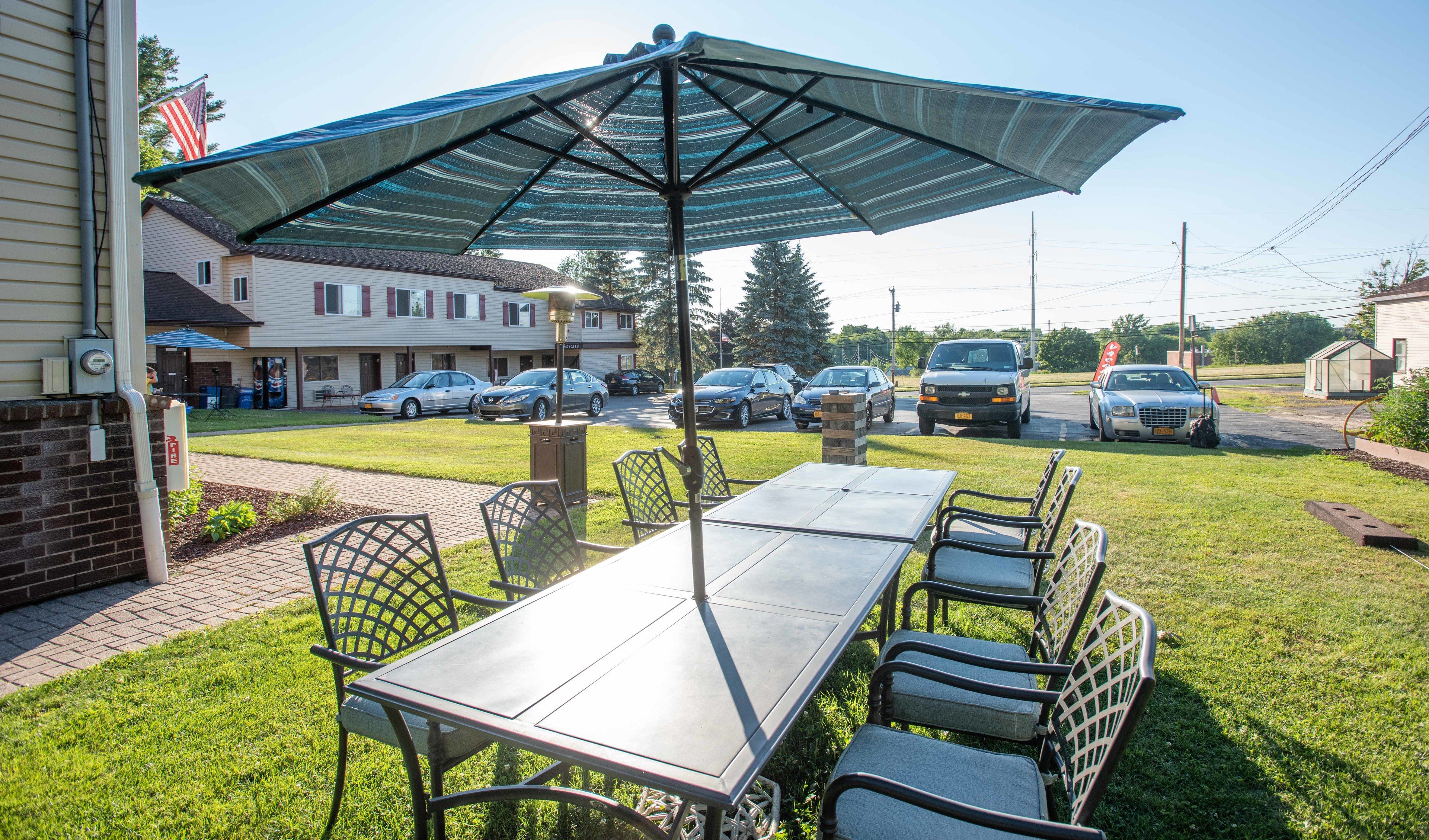 Eine Terrasse im Freien mit Tisch und Stühlen, einem großen Sonnenschirm und Blick auf parkende Autos und Häuser im Hintergrund.