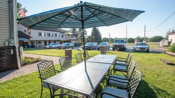 Eine Terrasse im Freien mit Tisch und Stühlen, einem großen Sonnenschirm und Blick auf parkende Autos und Häuser im Hintergrund.