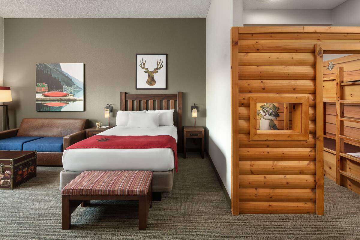 A hotel room with a wooden bed, a wooden bench, a suitcase, a lamp, and a framed picture of a deer on the wall.