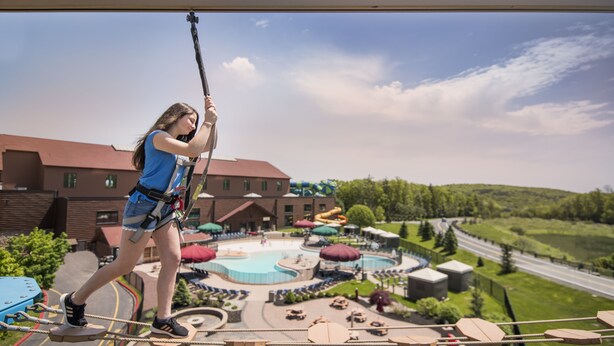A person on a zip line, overlooking a resort area with pools, lounge chairs, and a large building. - Great Wolf Lodge Concord (Concord)