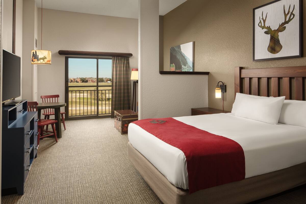 A hotel room with a large bed, a desk with a chair, a television, and a balcony with a view.