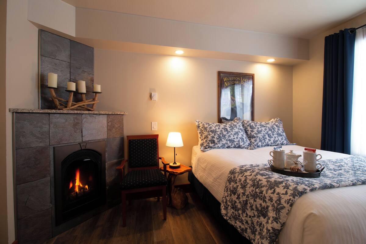 A hotel room with a fireplace, a bed with floral bedding, a nightstand with a lamp, and a tray with cups and a teapot.