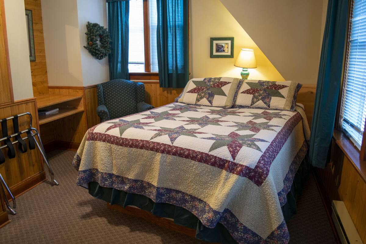 A neatly made bed with a star-patterned quilt, a chair, a wooden desk, and a lamp on the bedside table.