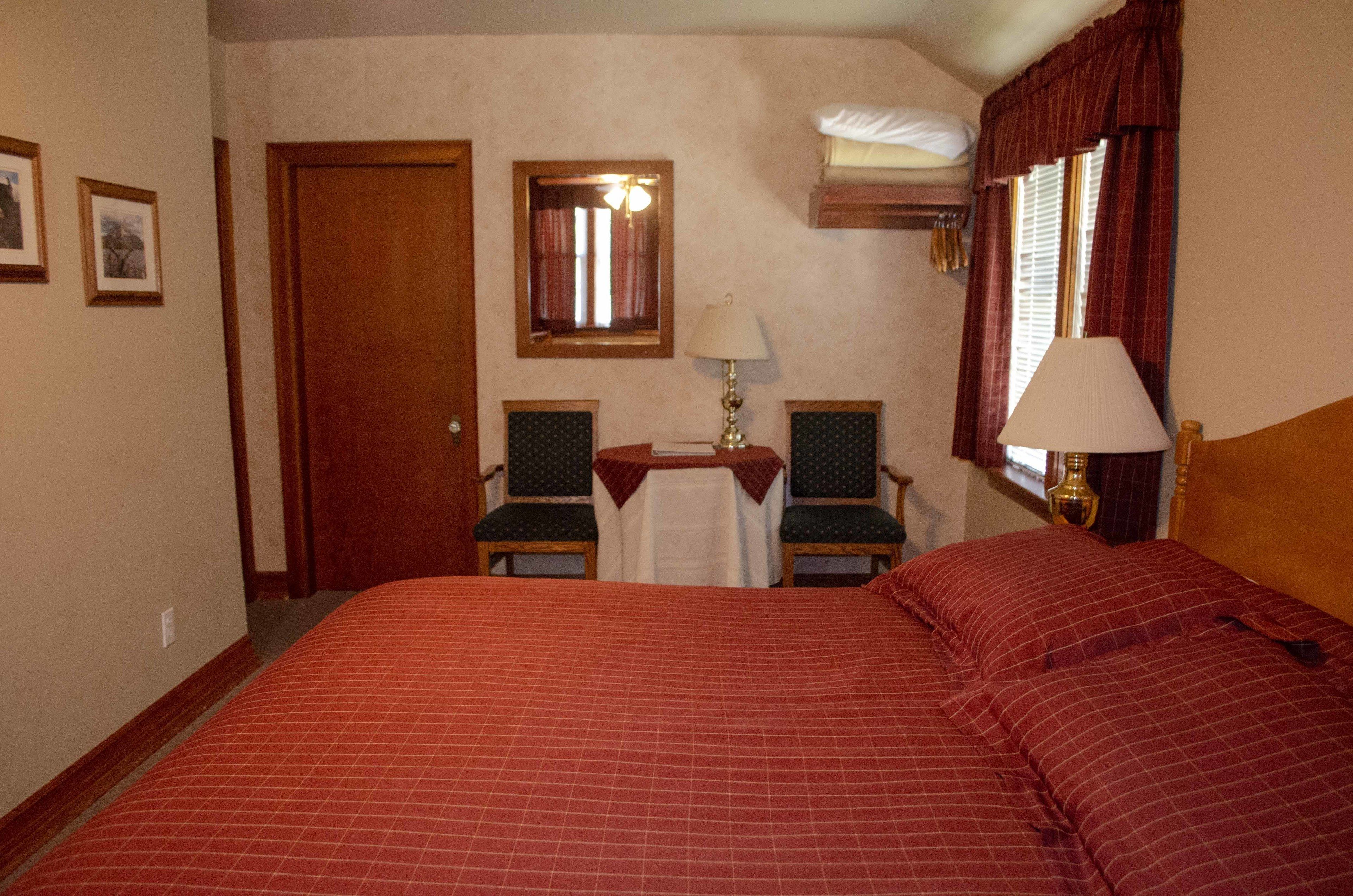 Une chambre d’hôtel avec un grand lit, deux chaises, une petite table avec une lampe, un miroir et une fenêtre avec des rideaux.